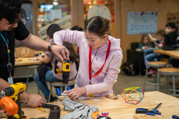 A teenage girl with safety glasses using a drill to create a project in the Tinkering Workshop at the Scott Family Amazeum.